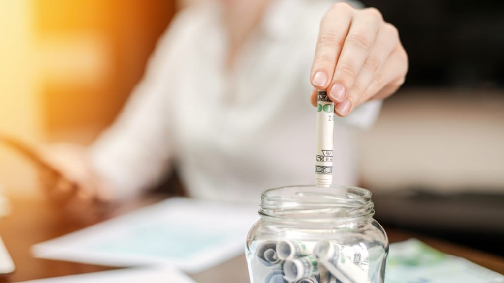 woman dropping banknotes into jar with rolled banknotes on the table papers on the table 1920x1080